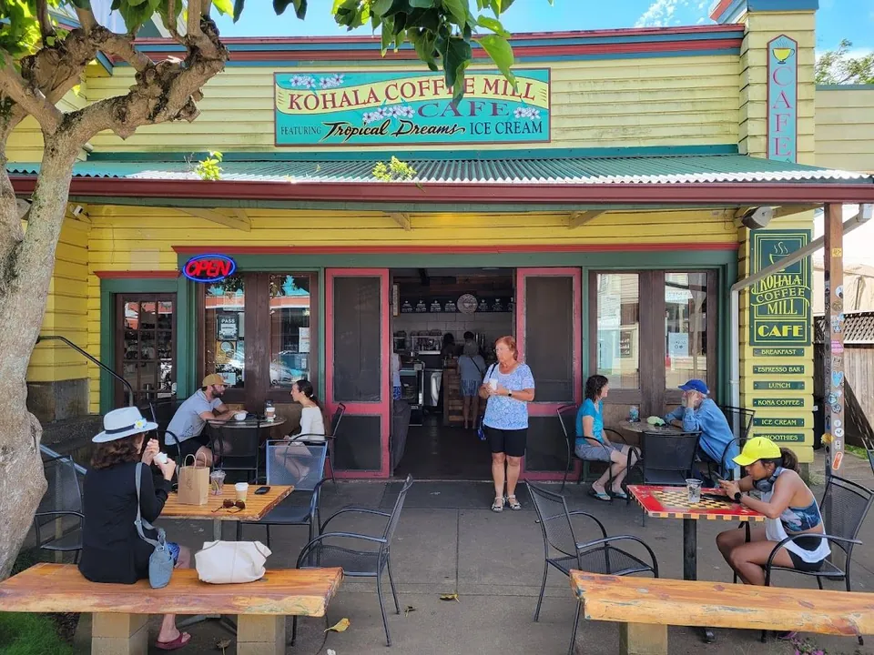 People sit at tables and one person stands outside the yellow Kohala Coffee Mill Cafe.