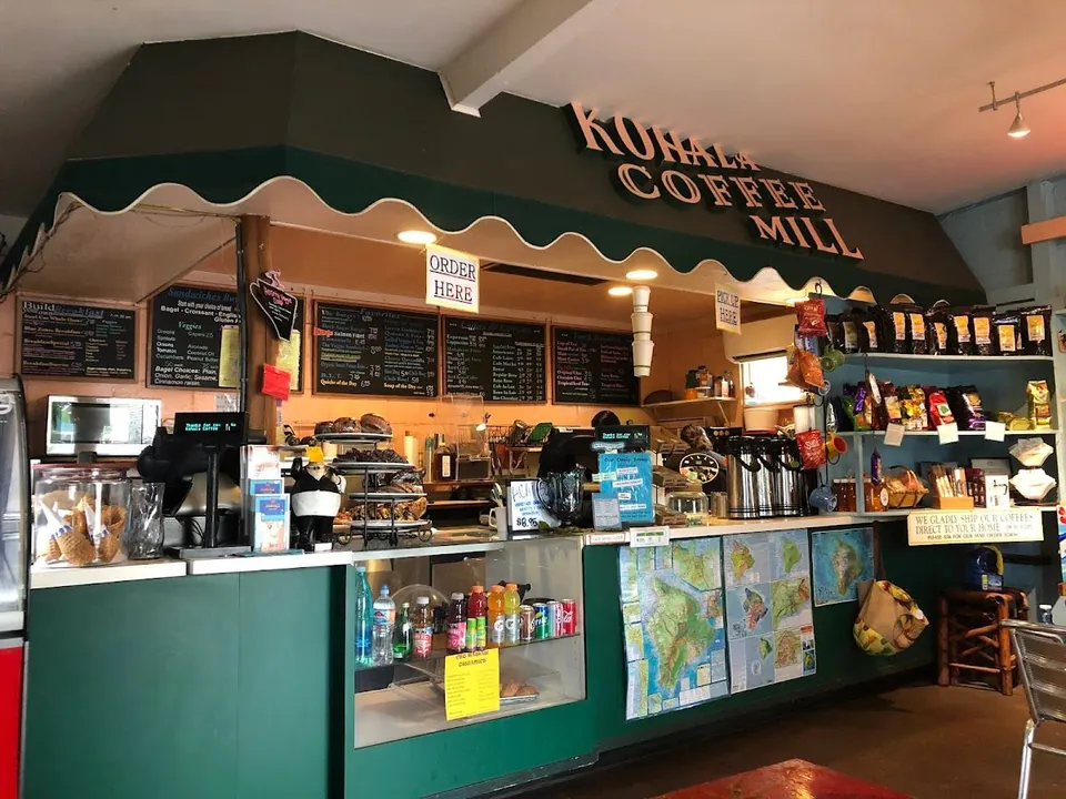 Kohala Coffee Mill counter under a green awning, with menu boards, a pastry case, and coffee bags on shelves.