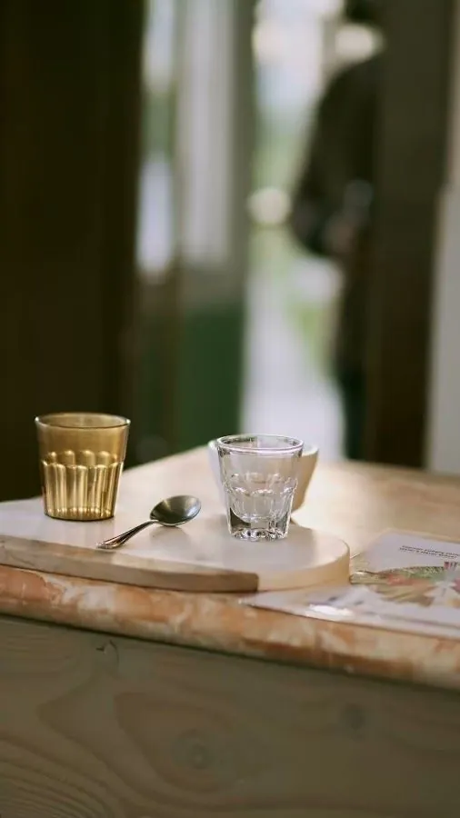 Amber and clear glasses, a spoon, and papers sit on a stone counter while a person stands blurred in the doorway.