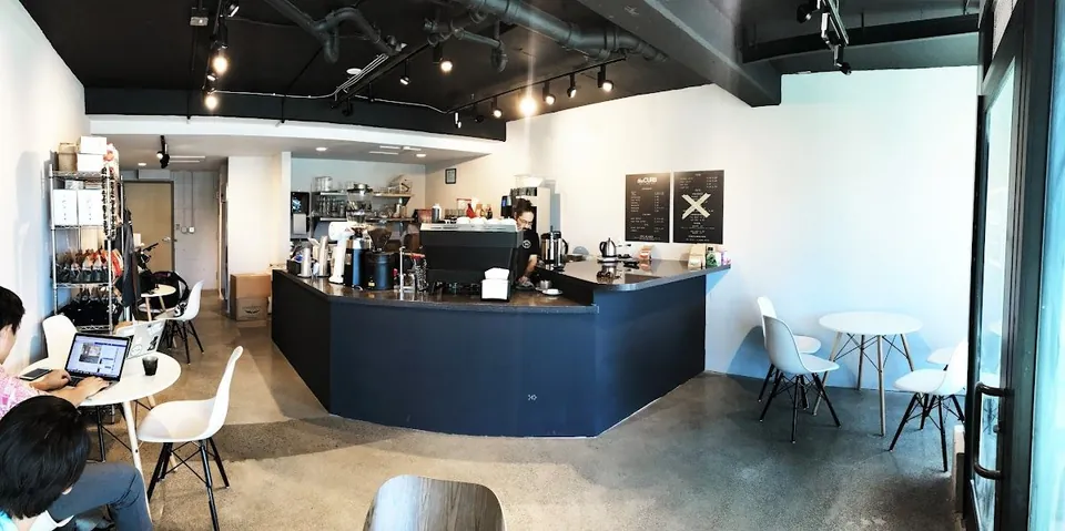 Cafe interior with a dark service counter, coffee equipment, wall menus, and people seated at tables.