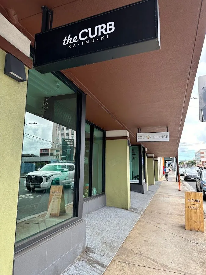 Storefront under a walkway awning, black sign the CURB KAI·MU·KI, wooden board, and a Color Lounge sign beyond.