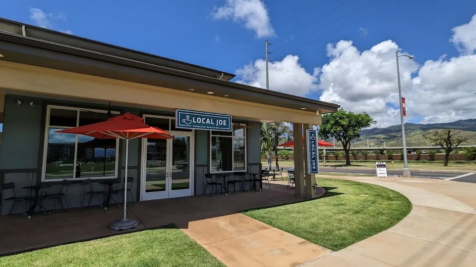 Storefront of a single-story shop with a covered patio, outdoor tables, a red umbrella, and a Local Joe sign.