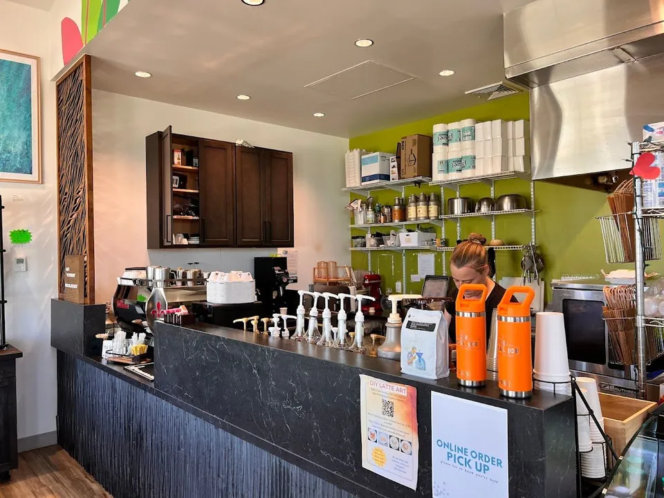 A worker looks down behind a dark counter with an espresso machine, syrup pumps, and pickup signs.
