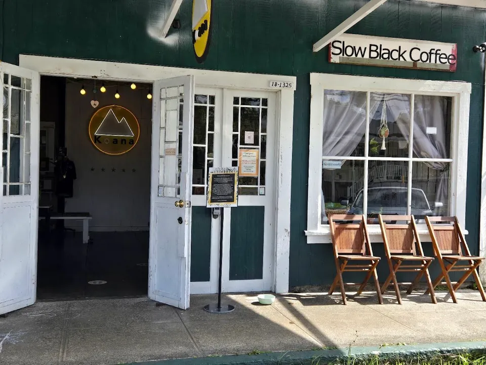 Dark green shop front with open white doors, a Slow Black Coffee sign, and three wooden chairs.