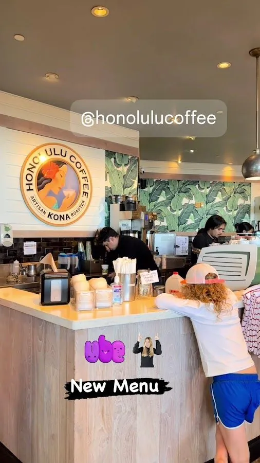 A customer in a white cap and blue shorts leans on the wood counter while staff work behind it at Honolulu Coffee.