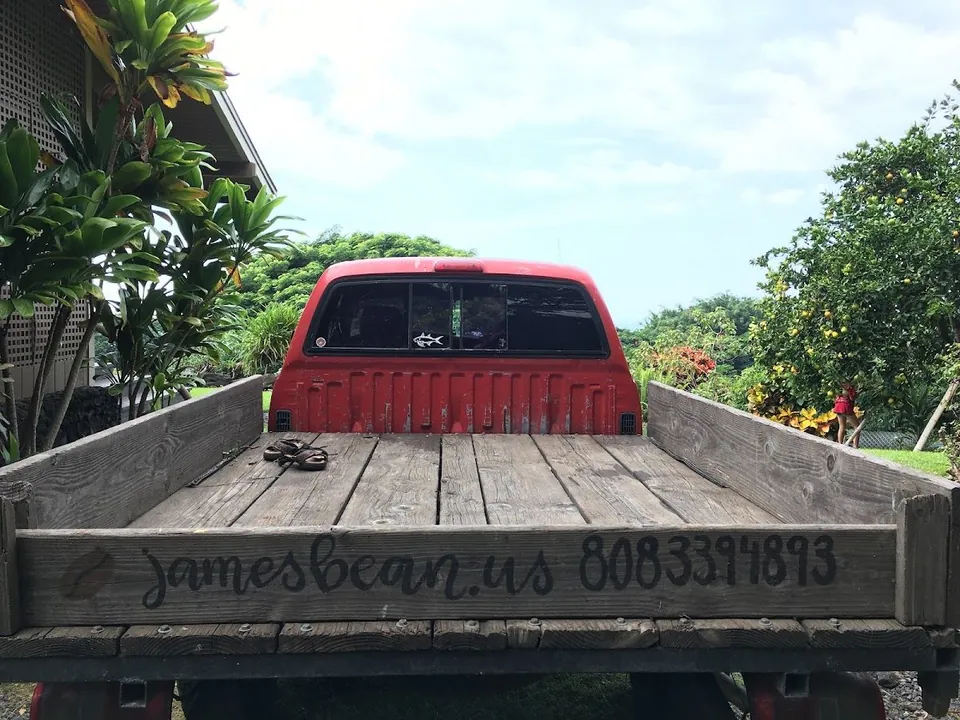 Red pickup with a wooden bed painted jamesbean.us and 8083394893, parked by a house and fruit trees.