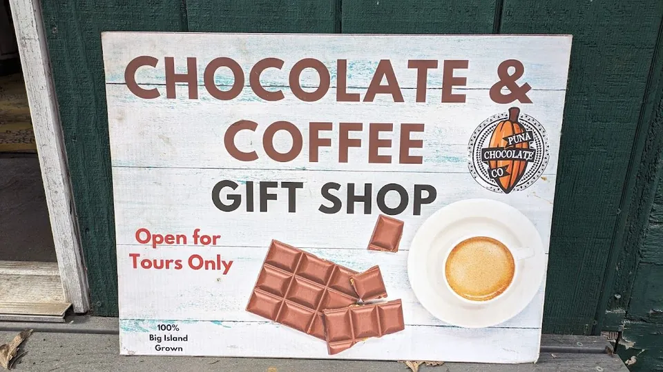 A painted Chocolate & Coffee Gift Shop sign, open for tours only, leans against a dark green wall.