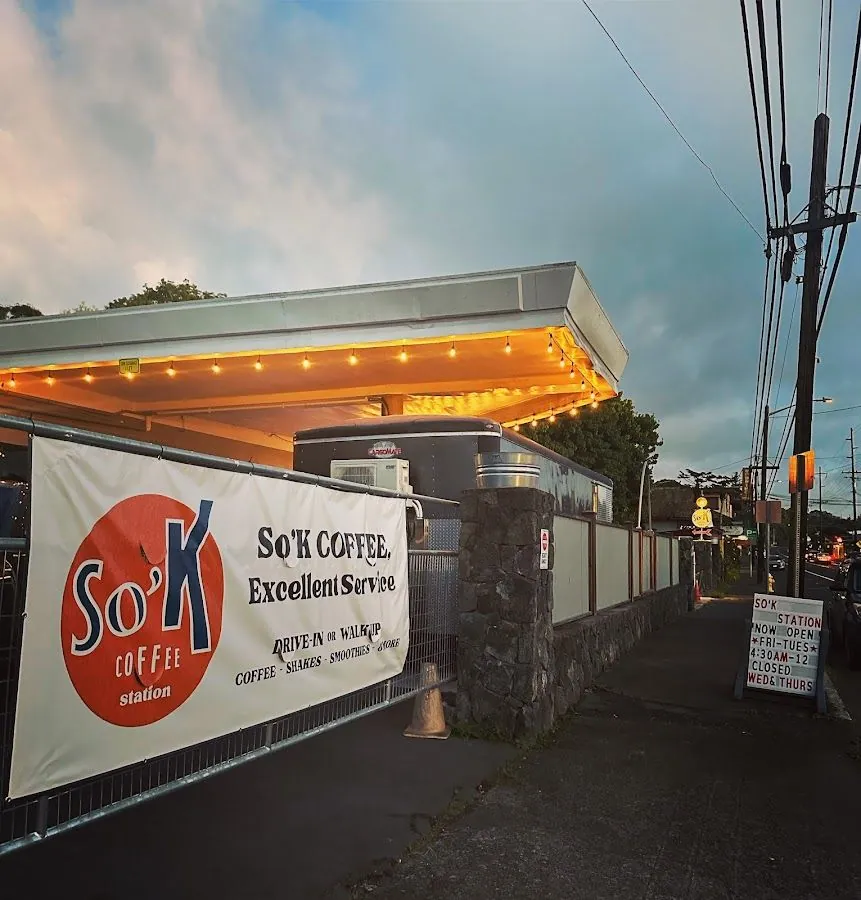 Dusk exterior of So'K Coffee Station with a lit canopy, fence banner, and sidewalk hours sign.