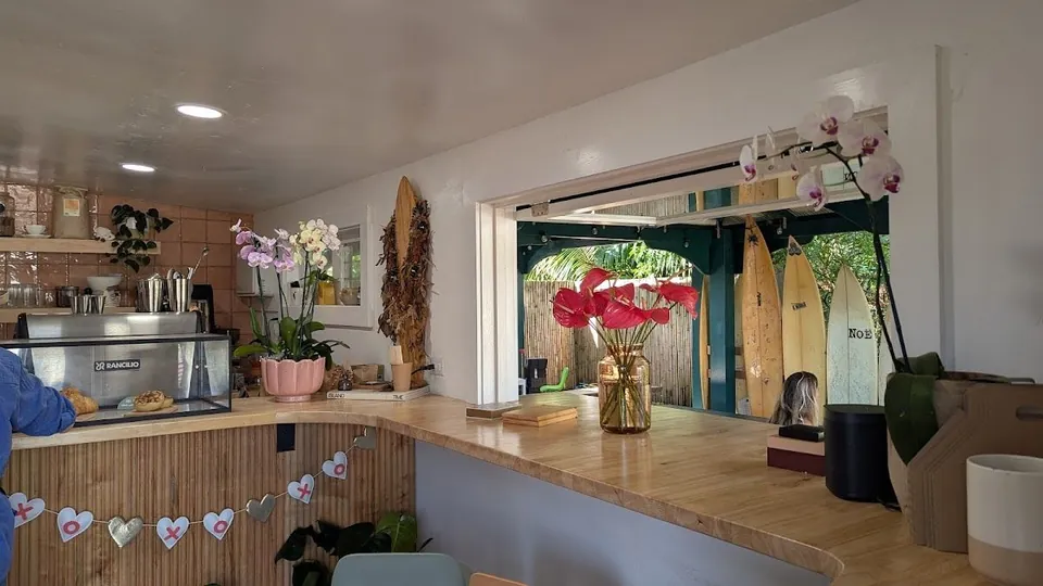 Cafe counter with an espresso machine, flowers, and an open window showing surfboards outside.