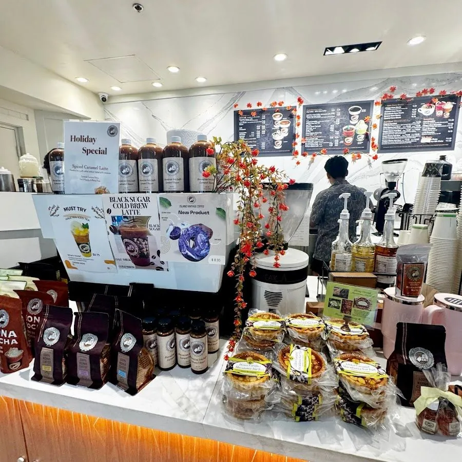 Cafe counter with coffee bags, drink bottles, packaged tarts, wall menus, and a worker at the bar.