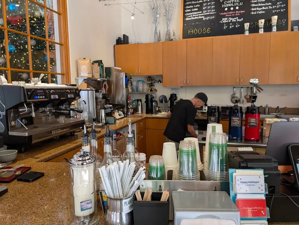 Coffee shop service counter with an espresso machine, cup stacks, syrup bottles, chalkboard menus, and a worker in black.