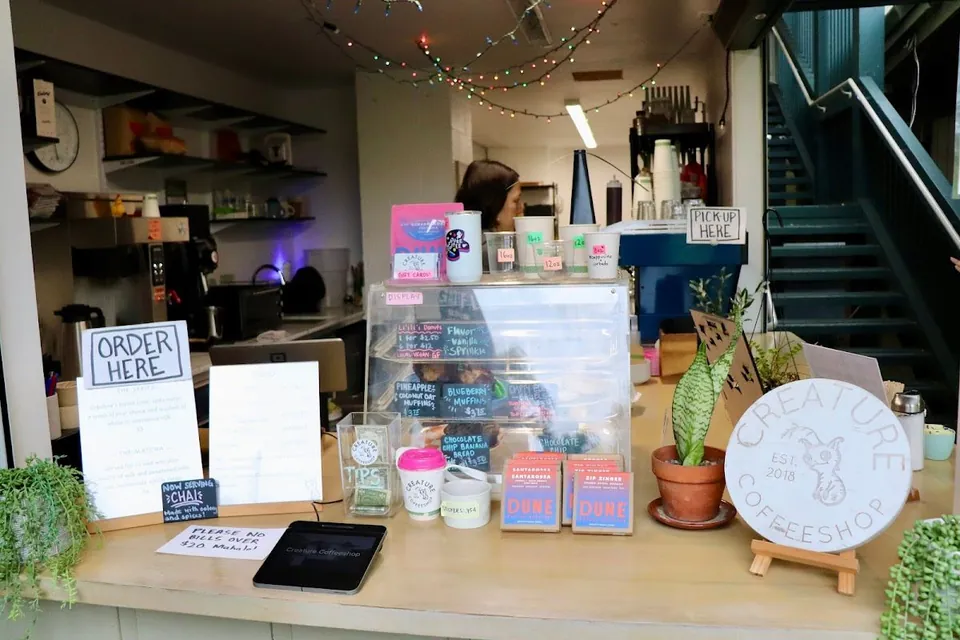 Cafe counter with order and pickup signs, a pastry case, plants, and a Creature Coffeeshop sign.
