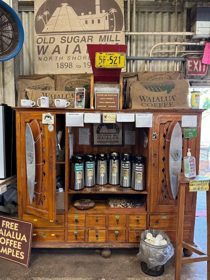 Wooden cabinet in a mill interior displays Waialua Coffee sacks, airpots, and sampling signs.