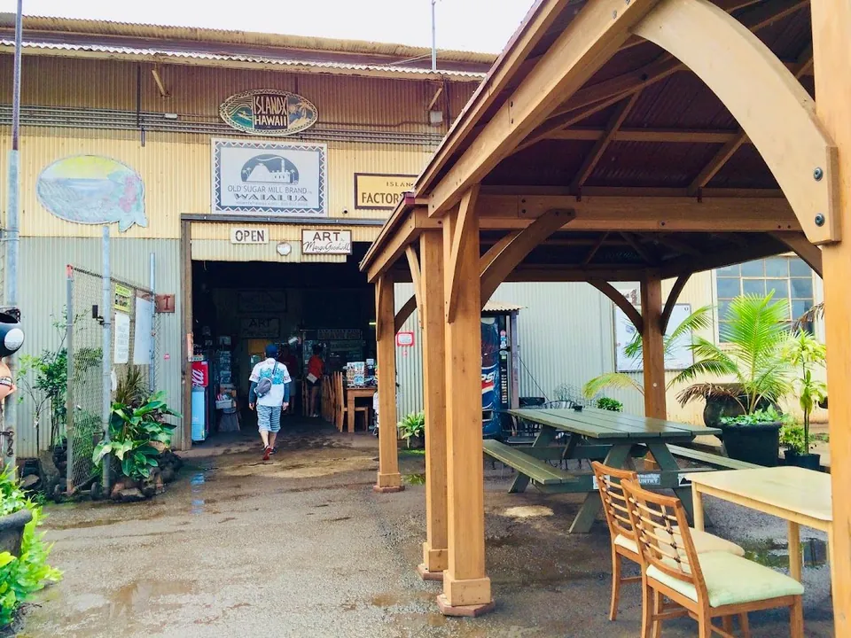A person walks into the open Island X Hawaii entrance beside a wooden pavilion and picnic tables.
