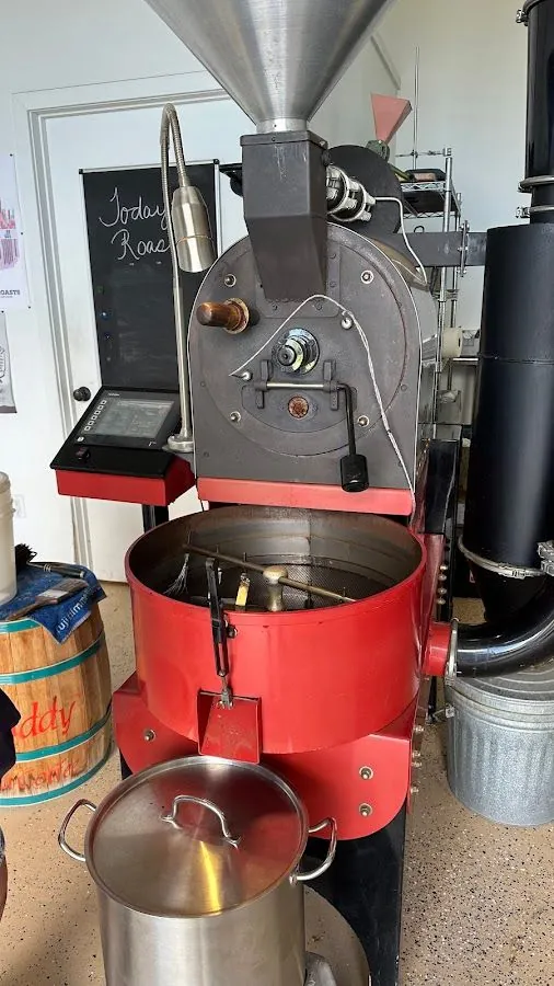 A red coffee roaster with an open cooling bin, hopper, control screen, and black exhaust duct.