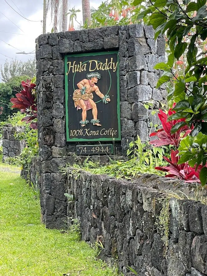 A stone pillar holds a Hula Daddy sign with a painted hula dancer and the words 100% Kona Coffee.