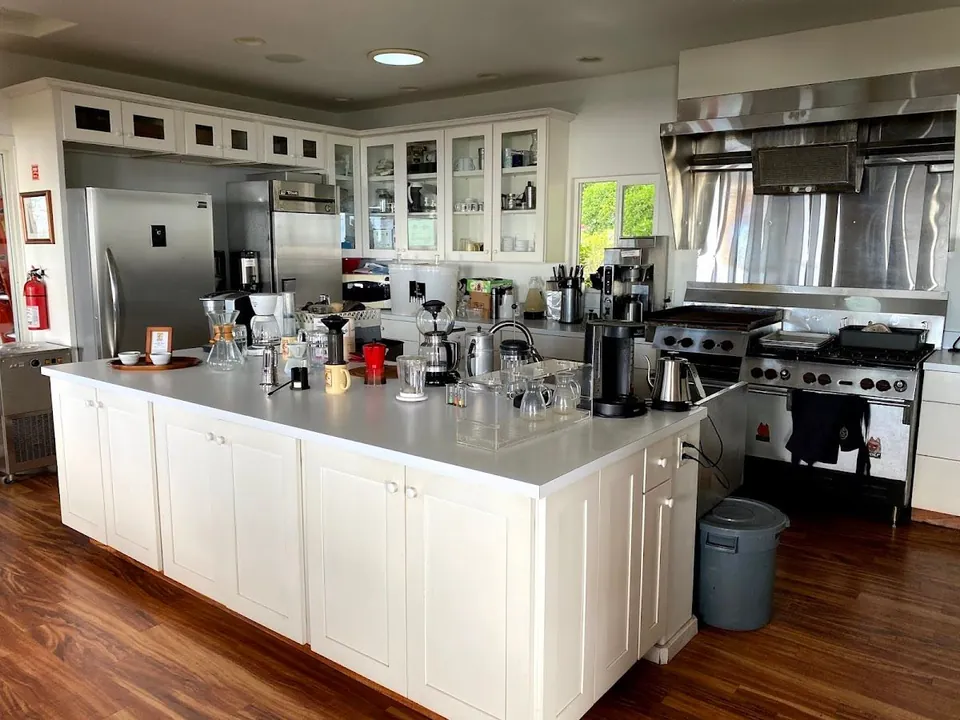 Commercial kitchen with a white island of coffee brewers, carafes, and kettles, plus stainless refrigerators and a range.
