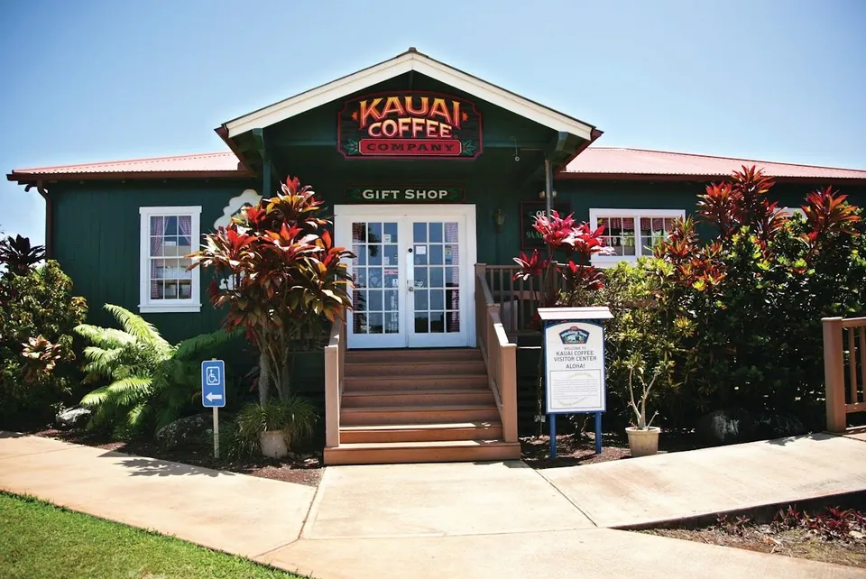 Green Kauai Coffee Company gift shop and visitor center with a red roof, front stairs, and tropical plants.