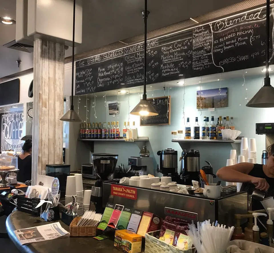 Cafe service counter under a large chalkboard menu, with espresso equipment, cups, syrup bottles, and two workers.