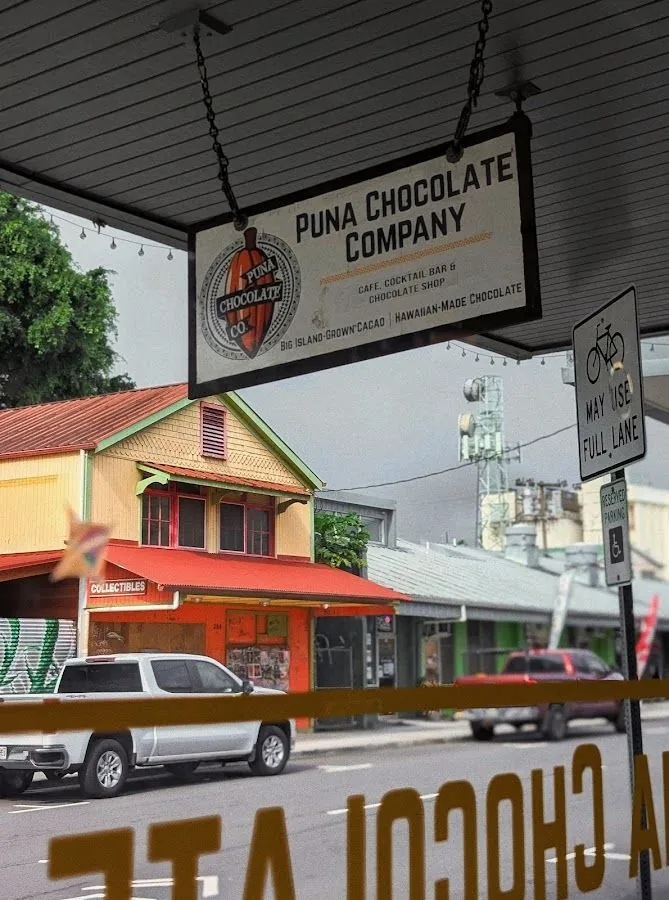 Puna Chocolate Company sign hangs from a covered walkway above a street of shops and parked trucks.