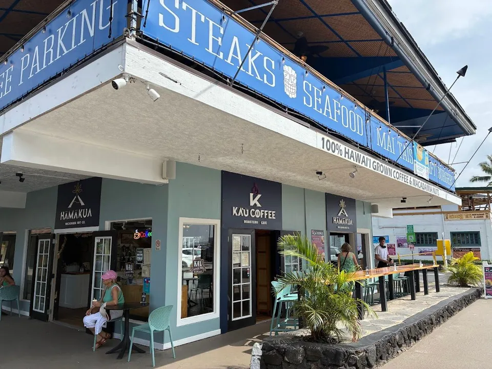 Storefront with Kaʻū Coffee and Hamakua signs, outdoor tables, and people seated and standing.