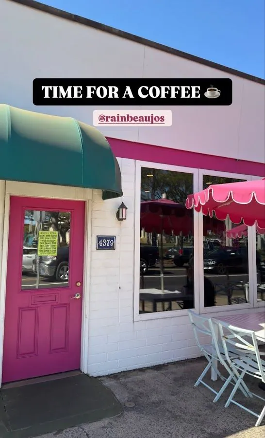 White shop front with a pink door, teal awning, wall number 4379, pink umbrellas, and patio chairs.