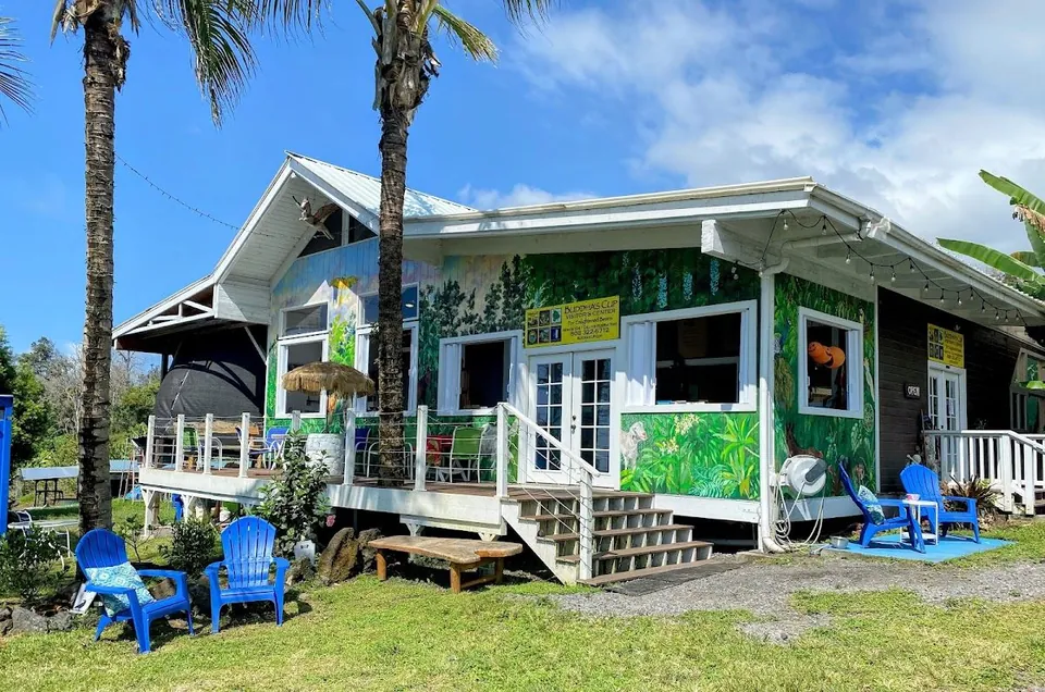 White farm building with a green mural, palm trees, and blue chairs on the lawn.