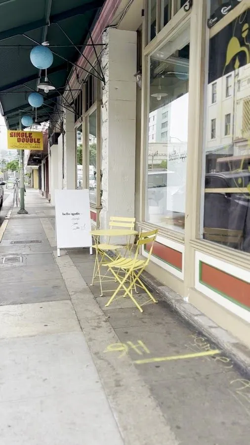 Yellow metal chairs, a small table, and a Hello Again sign stand on the sidewalk outside a shop window.