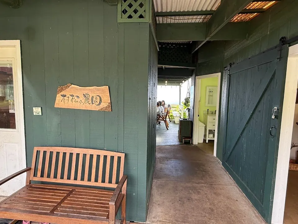 Covered dark-green porch with a wooden bench, a sign reading 村松の農園, a sliding barn door, and people seated on a far deck.