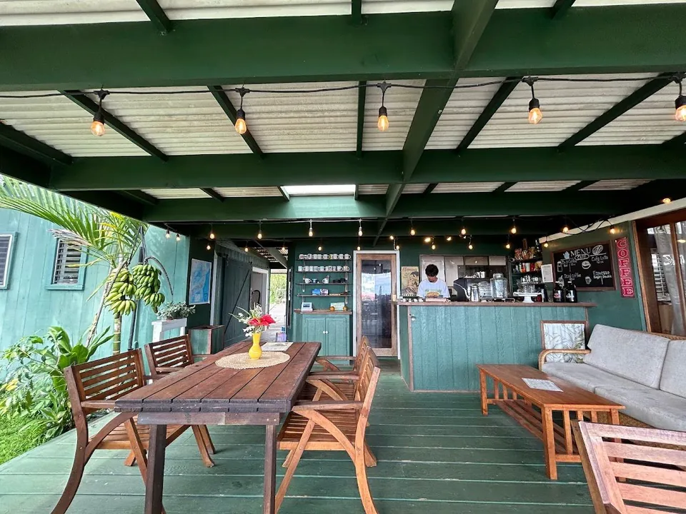 Covered green cafe porch with wooden tables, string lights, a service counter, and a person working behind it.