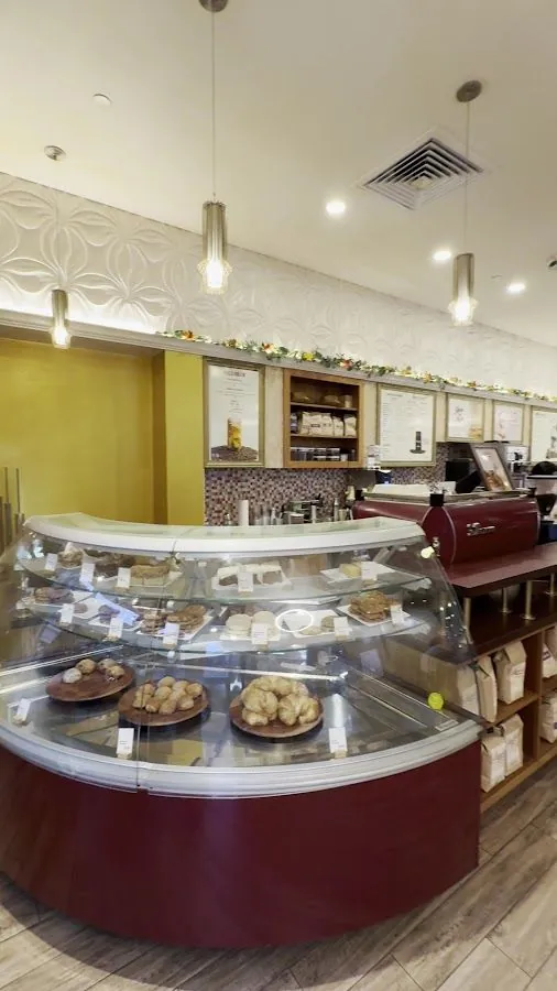 Cafe counter with a curved glass case of baked goods, a red espresso machine, coffee bags, and wall menus.