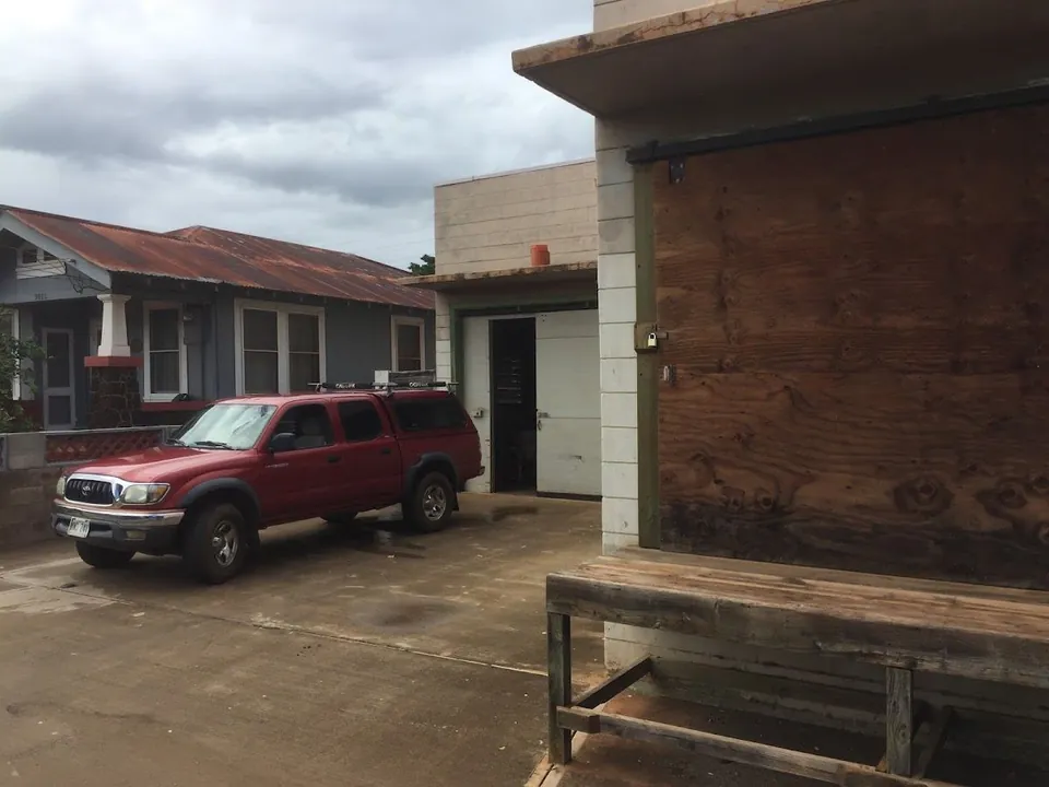 A red pickup truck sits on wet concrete by a gray house, an open garage, plywood boarding, and a wooden bench.