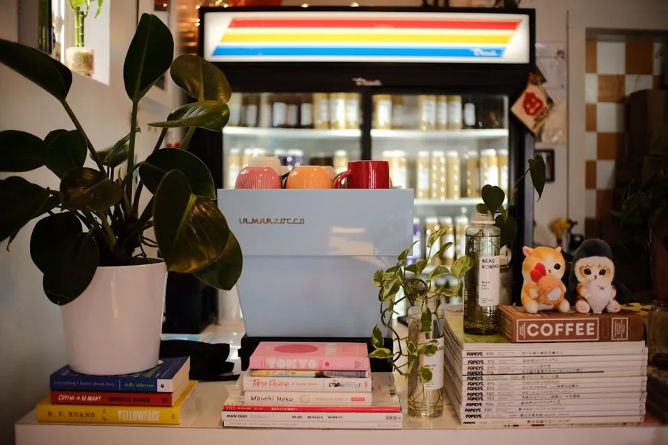 Light blue La Marzocco espresso machine on a cafe counter with plants, books, and cat plush toys.