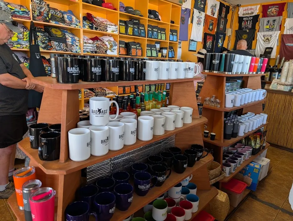 Wooden shop shelves hold rows of mugs, tumblers, coffee bags, and shirts, with people standing nearby.