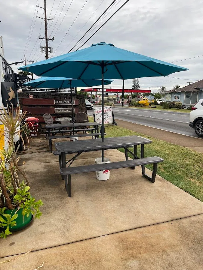 Outdoor picnic tables under blue umbrellas beside a road, with business signs on a stone wall.