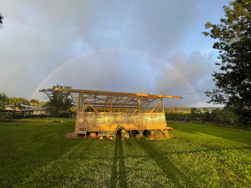 An open wooden shed with a corrugated metal roof and walls stands on a green lawn under a rainbow.