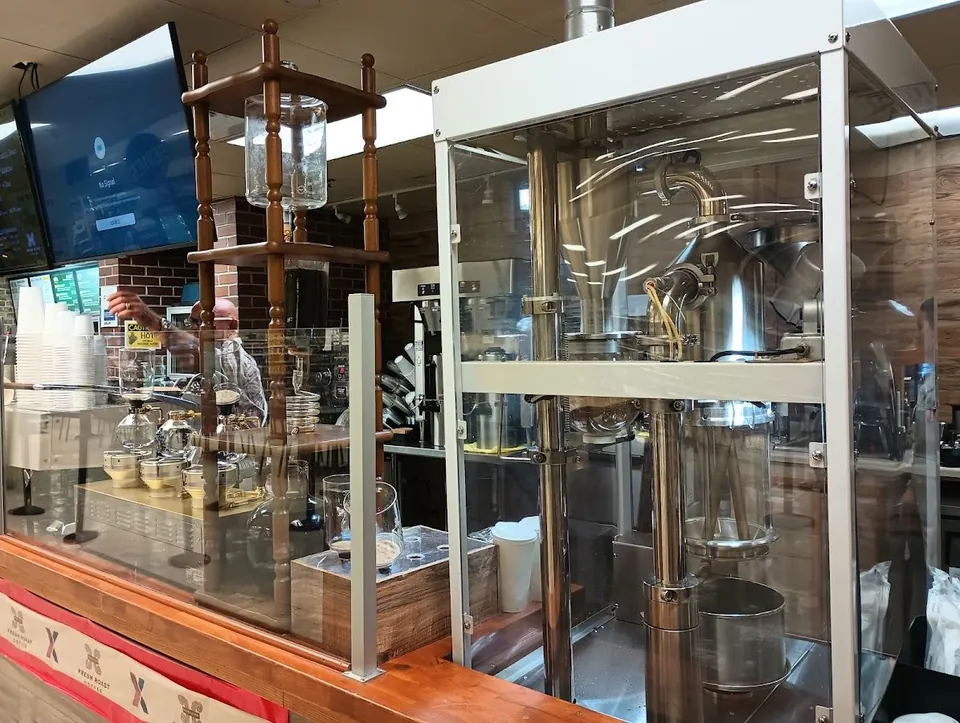 Coffee counter with a glass-cased metal roaster, pour-over brewers, stacked cups, and a worker behind the bar.