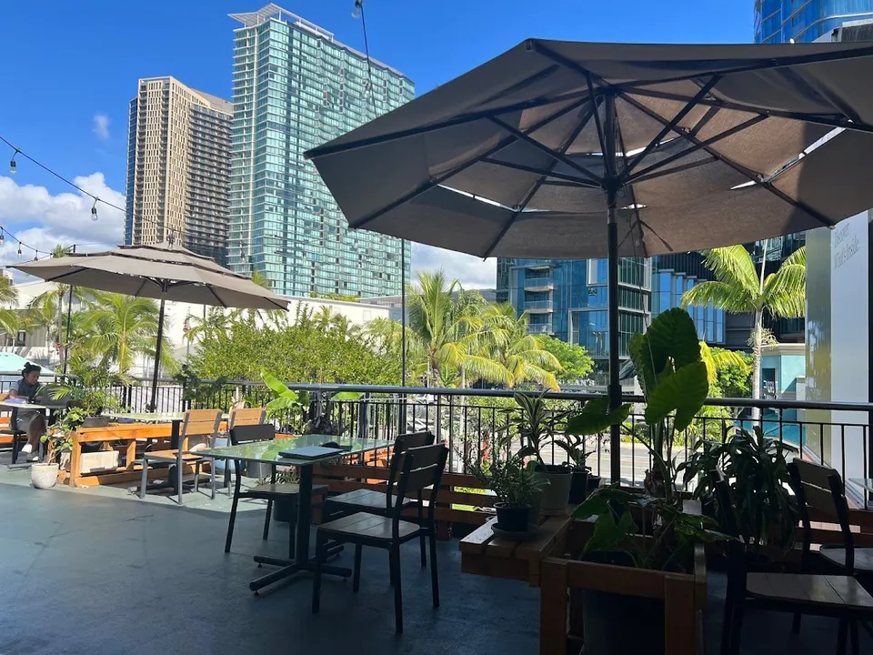 Open patio with umbrellas and tables; a person sits at the left and looks down, with towers beyond the railing.