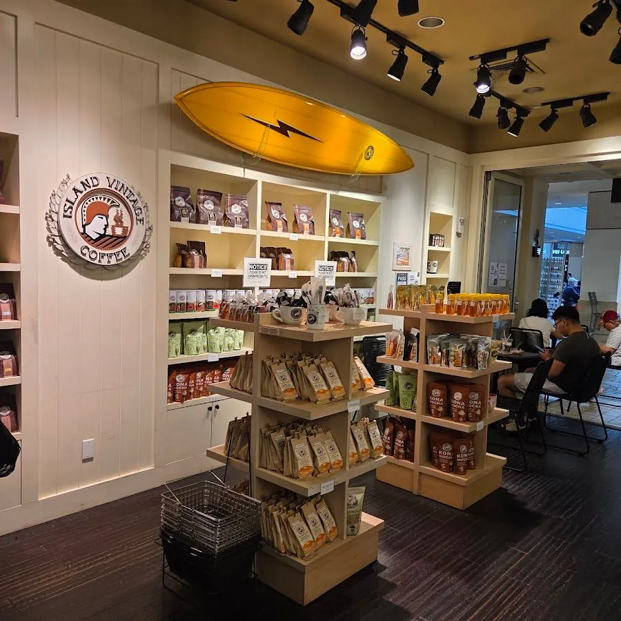 Shop interior with coffee-bag shelves, an Island Vintage Coffee sign, a yellow surfboard, and a seated person using a phone.