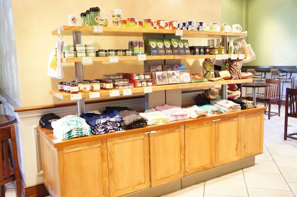 A light wood cabinet displays mugs, cups, jars, packaged goods, and folded shirts, with tables and chairs behind.