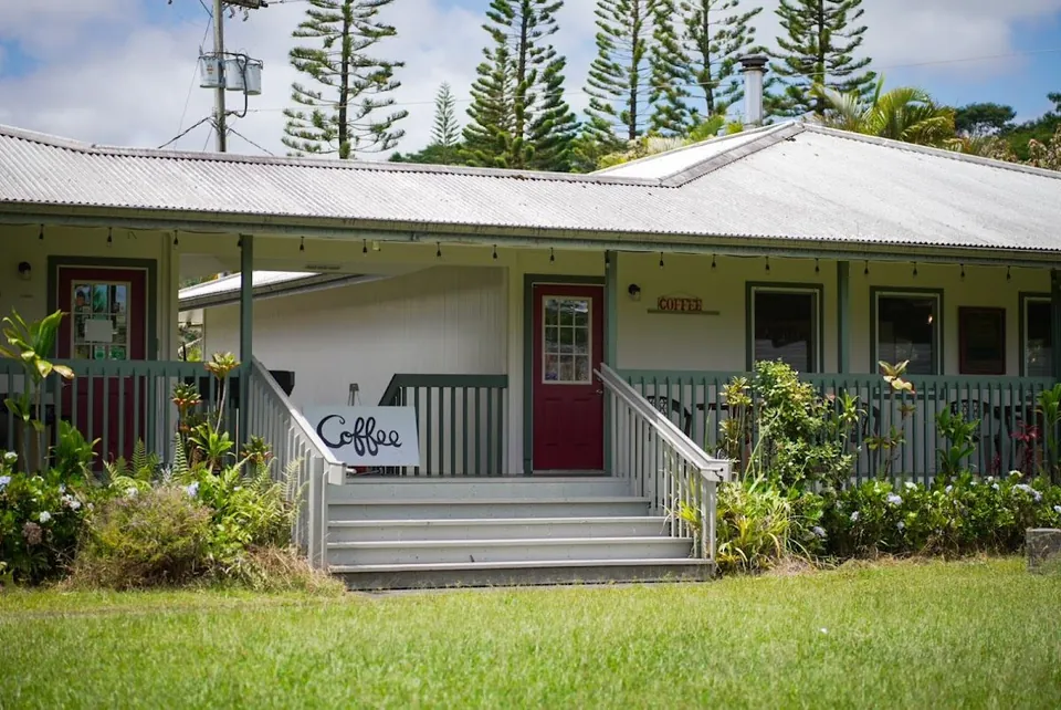 White single-story building with a metal roof, green porch, red door, steps, and a Coffee sign.