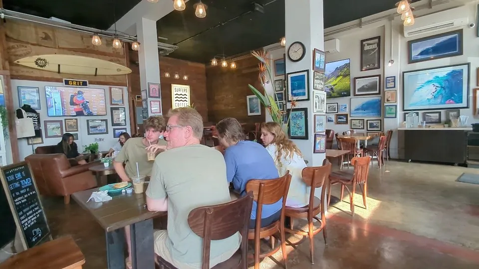 People sit at tables inside a coffee shop with a surfboard, pendant lights, and wave photos.