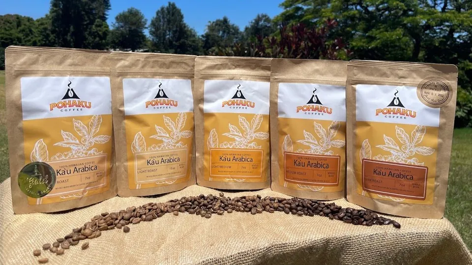 Five Pohaku Coffee bags of Kāu Arabica stand on burlap outdoors, with roasted beans scattered in front.