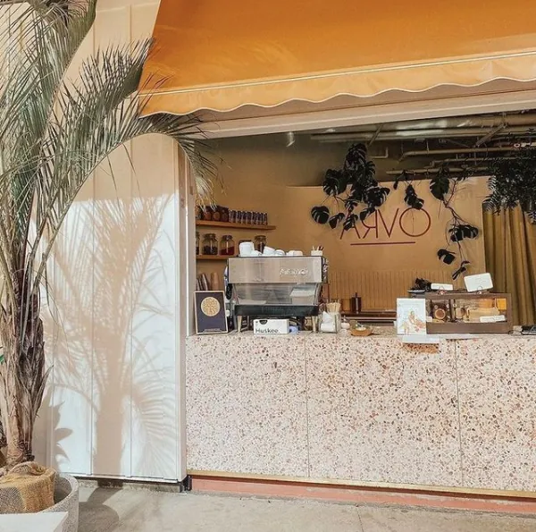 Cafe service window under an orange awning, with a speckled counter, espresso machine, hanging plants, and a palm outside.