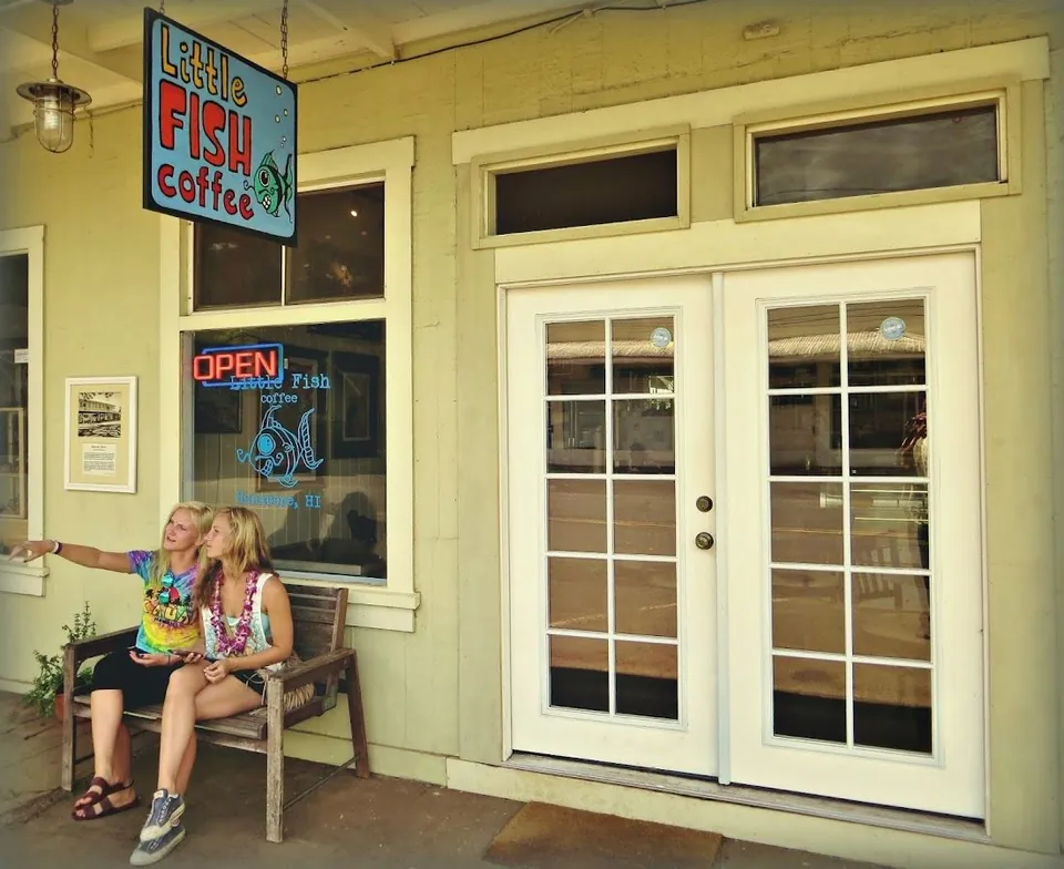 Two people sit on a bench outside Little Fish coffee, one pointing, beside white double doors.