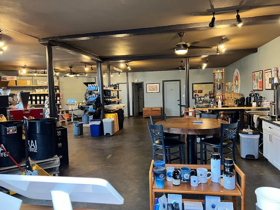 Coffee shop interior with a round table, mug cart, brewing equipment, and a person working at the left counter.