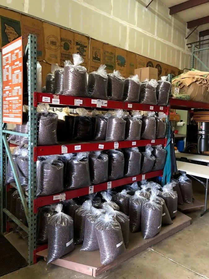 Clear plastic bags of dark coffee beans fill red metal shelves in a warehouse, with printed burlap sacks on the wall.