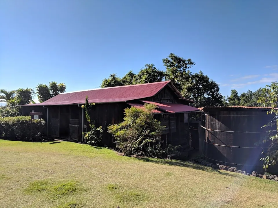 Dark wooden building with a red metal roof beside a large round wooden tank, on a lawn under trees and blue sky.