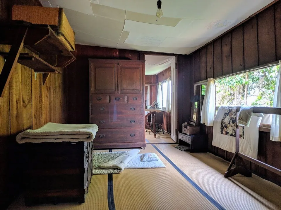 Wood-paneled room with tatami mats, a tall wooden chest, folded bedding, and white quilts by the windows.
