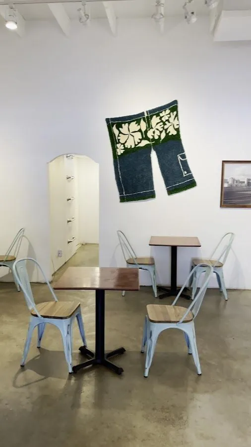 Seating area with pale blue metal chairs, wood tables, and a large shorts-shaped textile on a white wall.